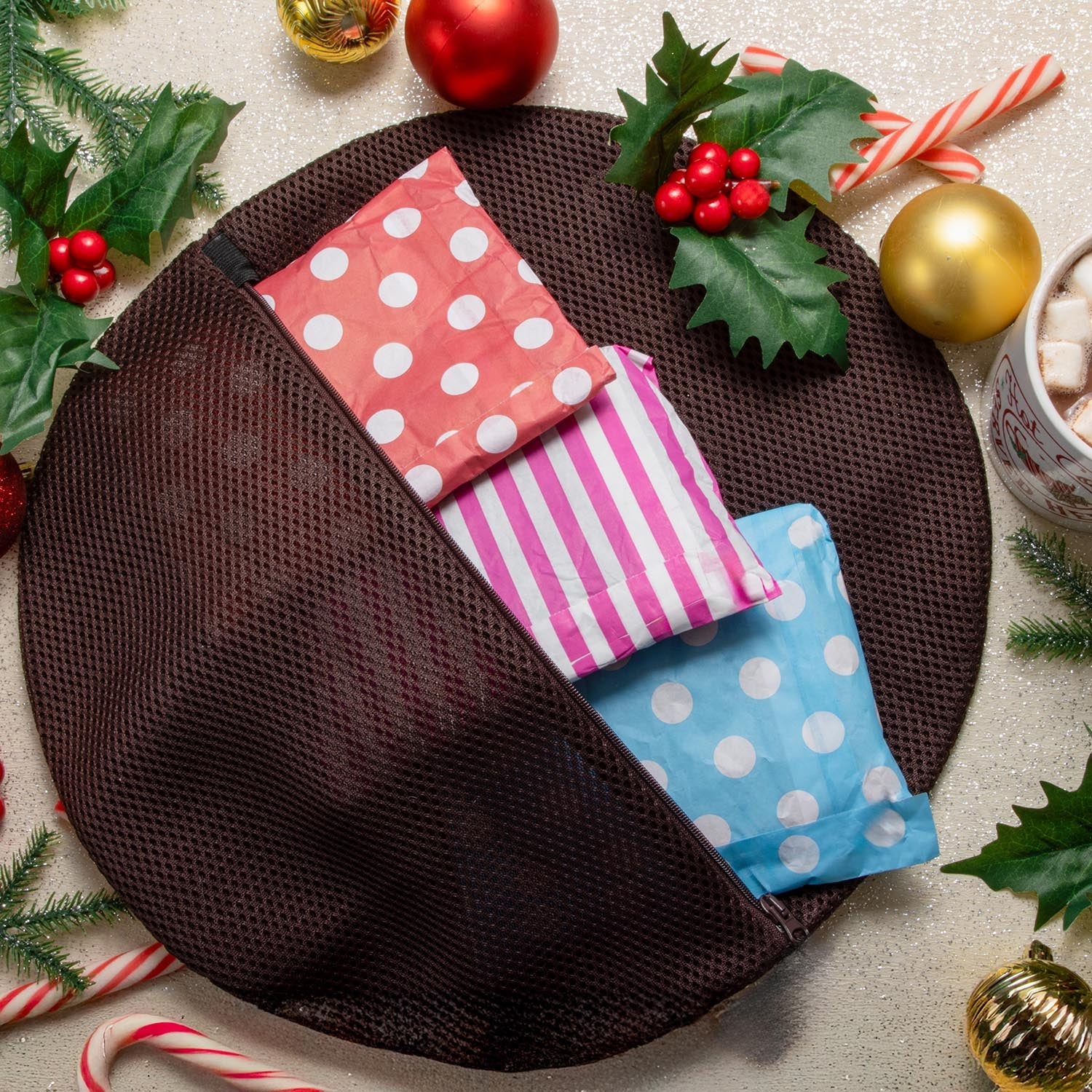 The reverse side of the Snagvent Christmas pudding washbag, showing the brown mesh fabric and zipper opening. Tucked inside are three pairs of tights in colorful paper packaging—red with white polka dots, pink and white stripes, and blue with white polka dots—set against a festive background with holly and baubles.